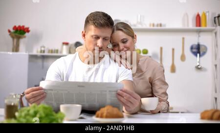 Giovane marito leggendo il giornale con la moglie holding tazza da caffè, mattina insieme Foto Stock