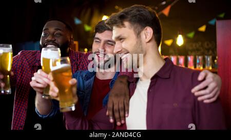 Ubriaco amici multietnica tintinnanti bicchieri da birra, trascorrendo il tempo in pub, party Foto Stock