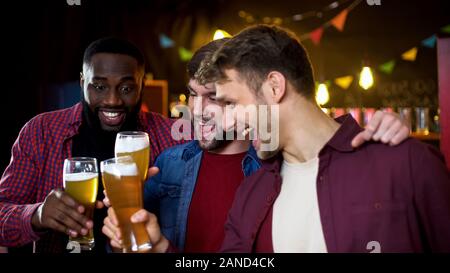 Multietnica gioiosa bachelors tintinnanti bicchieri da birra, guardando la partita di calcio in pub Foto Stock