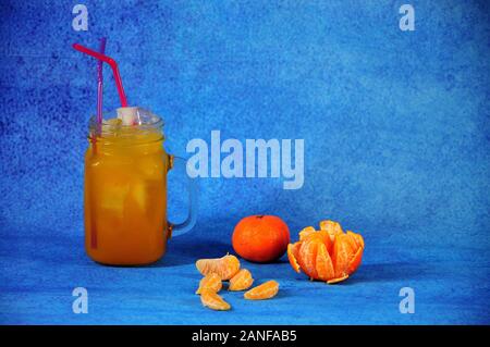 Tazza di vetro di succo di mandarino con ghiaccio e cannucce accanto a fette di frutta e tutto il mandarino su uno sfondo blu. Close-up. Foto Stock