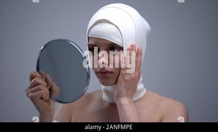 Signora in headwrap elastico alla ricerca dello specchio, sensazione di dolore dopo un intervento di chirurgia plastica Foto Stock