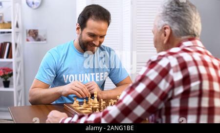 Di mezza età volontari maschi giocando a scacchi con uomo anziano in casa di cura, hobby Foto Stock