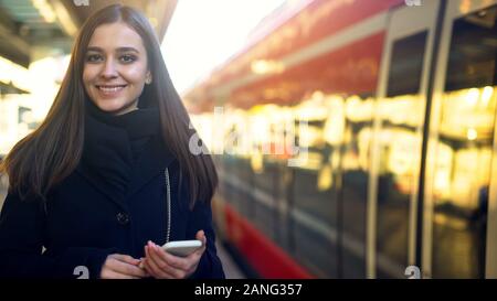 Donna con telefono sorridendo vicino stazione, rapido pagamento mobile per la tecnologia dei biglietti Foto Stock