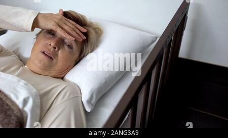 Sconvolto femmina senior giacente in letto di ospedale, toccando la fronte, sofferenza emicrania Foto Stock