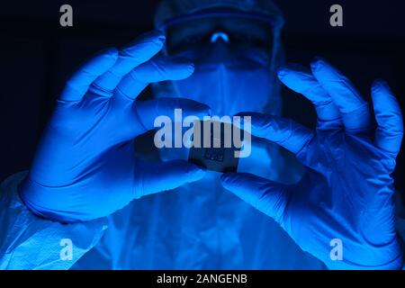 Un uomo con una tuta di protezione contiene un microchip nelle sue mani. Laboratorio di ricerca al lavoratore che indossa guanti e occhiali di sicurezza. Foto Stock