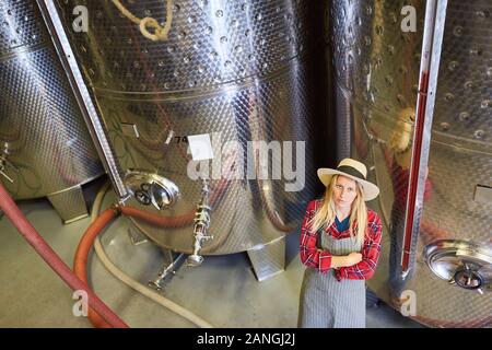 Giovane donna come un enologo partecipante nella parte anteriore di un serbatoio di fermentazione mentre l'imbottigliamento del vino Foto Stock