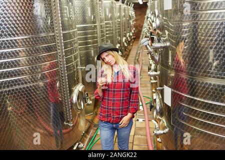 Giovane donna come un enologo nella parte anteriore del serbatoio di fermentazione in cantina con un bicchiere di vino rosso Foto Stock