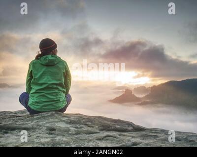 Viaggiare da soli donna escursionista si siede sul bordo della scogliera e godendovi l'alba guardando la valle e le montagne. Viaggiando uno stile di vita attivo concept Foto Stock