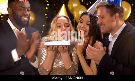 Felice bionda femmina torta di soffiaggio candele, amici congratularmi con donna compleanno Foto Stock