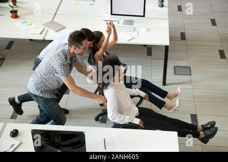 Emozionato diversi dipendenti divertirsi in ufficio a cavallo su sedie durante la pausa di lavoro, multirazziale millenaria colleghi ridere divertente al lavoro, pl Foto Stock