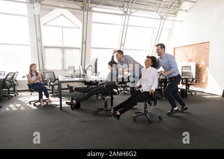 Emozionato millenario lavoratori diversi divertirsi in office racing su sedie insieme, felice multirazziale dipendenti impegnati in attività di team building, laughin Foto Stock