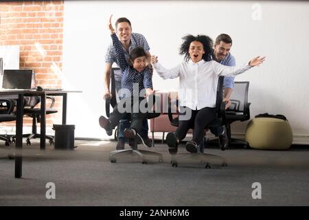 Emozionato multirazziale i dipendenti giovani divertirsi in ufficio a cavallo su sedie vincendo la gara, felice diverse intrattenere i lavoratori sul luogo di lavoro si impegnano a ridere Foto Stock
