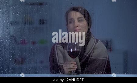 Malinconici donna in coperta si beve vino dietro la finestra piovosa, fredda sera Foto Stock