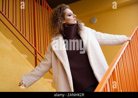 Una ragazza con il rosso dei capelli ricci in camice bianco pone sul giallo arancione parcheggio scale. City Style - Urban Foto Stock