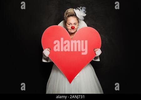 Donna felice clown mostra rosso cuore vuoto banner su sfondo nero Foto Stock