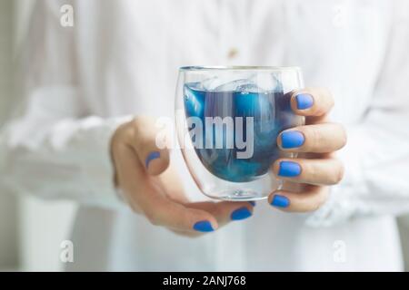 Matcha blu bevanda fredda con ghiaccio in un bicchiere di vetro nelle mani di una donna con manicure blu. Colore dell'anno 2020 trend Foto Stock