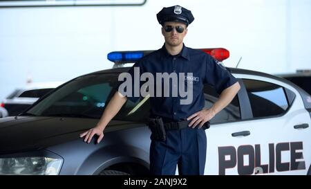Coraggioso poliziotto in auto di servizio e occhiali da sole in posa sulla fotocamera in prossimità di auto di pattuglia Foto Stock
