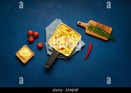 Tavolo per la colazione con uova strapazzate in padella con formaggio, jalapenos e pomodori sul classico sfondo blu. Lay piatto di uova strapazzate. Frittata cremosa Foto Stock