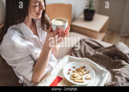 Bella giovane donna hanno una romantica colazione in mattina accogliente bed. Tazza di caffè e la parola amore da biscotti dolci. Sorpresa il giorno di San Valentino Foto Stock