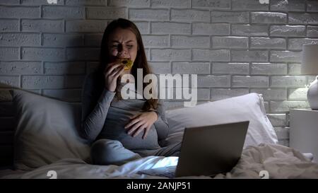 Giovane donna incinta di piangere nella parte anteriore del computer portatile, mangiare ciambella per superare lo stress Foto Stock