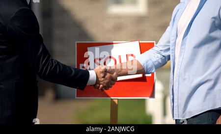 Agente immobiliare e il nuovo proprietario di casa stringono le mani nella parte anteriore del cartello venduto Foto Stock