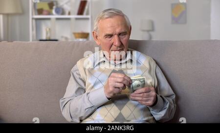 Premuto vecchio seduto sul divano di contenimento di banconote di dollari, insicurezza sociale Foto Stock