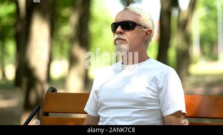 Vecchio senior in occhiali da sole seduto sulla panchina nel parco, proteggere gli occhi dai raggi del sole Foto Stock