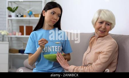 Sconvolto donna anziana che rifiuta di mangiare i fiocchi d'avena offerti da volontario, casa di cura Foto Stock