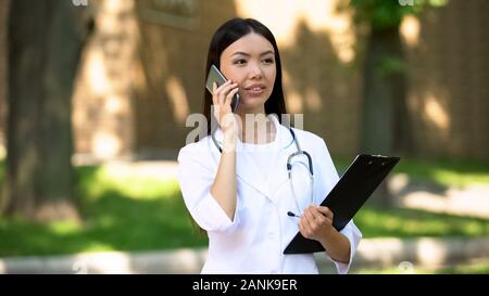 Piuttosto medico parlando con paziente sullo smartphone in ospedale park, 24h Foto Stock