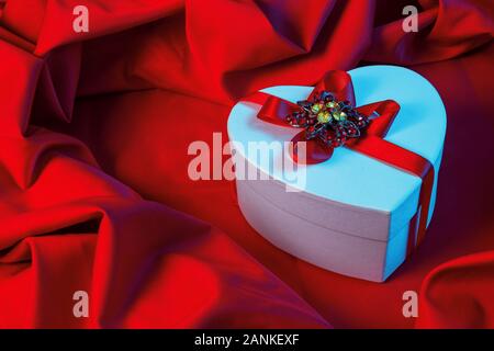 Valentines greeting card in luce blu. bianco scatola di cartone a forma di cuore avvolto in nastro giaceva su un panno rosso che riprende la forma del presente p Foto Stock