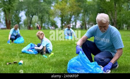 Senior man picking up litter nel parco famiglia insieme sorridente fotocamera, Earth care Foto Stock