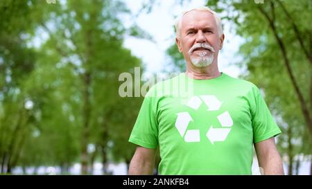 Grave il vecchio uomo che indossa la t-shirt con segno di riciclaggio, guardando la fotocamera all'aperto Foto Stock