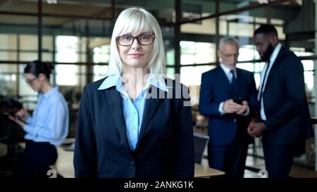 Business di successo signora sorridente in telecamera, colleghi sullo sfondo, l'occupazione Foto Stock