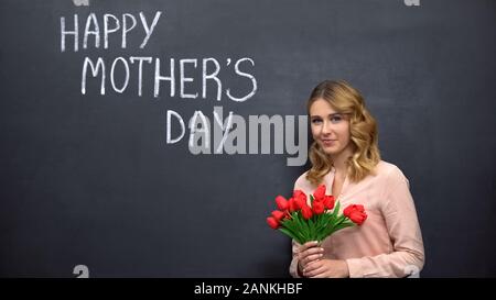 Donna con fiori in piedi vicino a firma felice festa della mamma, congratulazioni Foto Stock