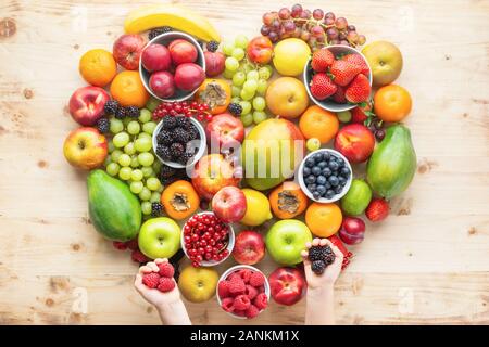 Bambino la mano di cuore di contenimento realizzato da sani frutti arcobaleno, Fragole Lamponi arance Prugne Mele Kiwi uve mirtilli mango papaya sulla tavola di legno, vista dall'alto, copia di spazio, il fuoco selettivo Foto Stock