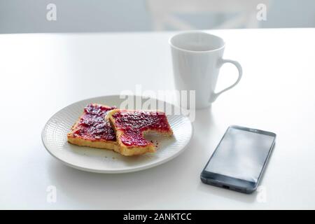 La colazione è a tavola. Una tazza di caffè e toast con marmellata di ciliegie accanto al telefono. Foto Stock