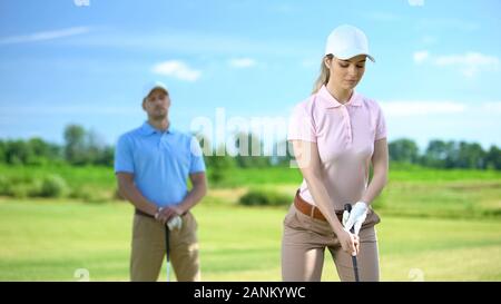 Pullman maschio guardando la ragazza golfista con club che mira a colpire la sfera in corso, la formazione Foto Stock