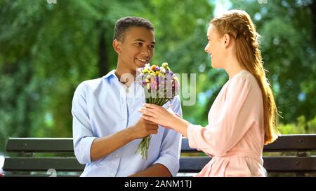 Sorridente mixed-gara teen boy presentando cara ragazza fiori, prima data in posizione di parcheggio Foto Stock