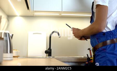 Tuttofare in uniforme controllo rubinetto da cucina, la digitazione di relazione nel tablet, idrosanitari Foto Stock