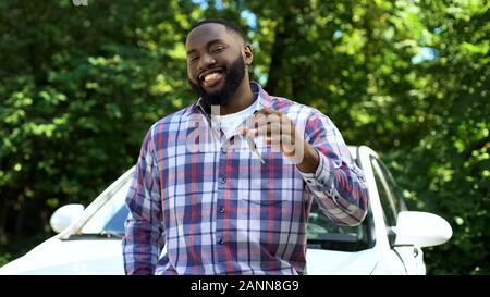 Sorridente afro-americano che mostra le chiavi dell'auto appoggiata nuova automobile, servizio di noleggio Foto Stock