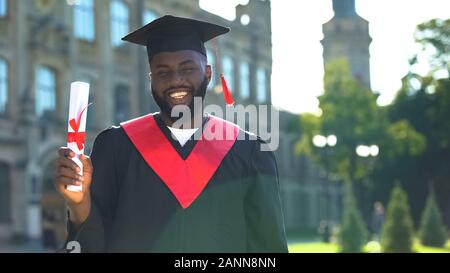 Emozionato afro-studente americano in abito di graduazione holding diploma guardando la fotocamera Foto Stock