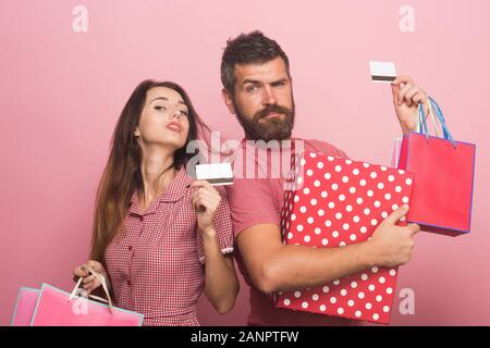Ragazzo con la barba e lady fare shopping. I soldi di spesa e di concetto di shopping. Uomo con grave di fronte si erge nei pressi di giovane donna. Matura in amore detiene il big box e bustine che mostra le carte di credito su sfondo rosa Foto Stock