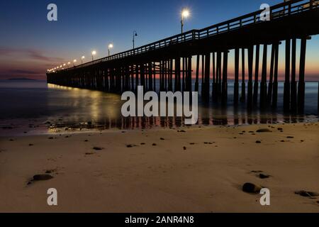 Ventura Pier al tramonto, Ventura, California. Sabbia e rocce in primo piano, acqua rientrante nella bassa marea. Lampade su pier; colorato sky in background. Foto Stock