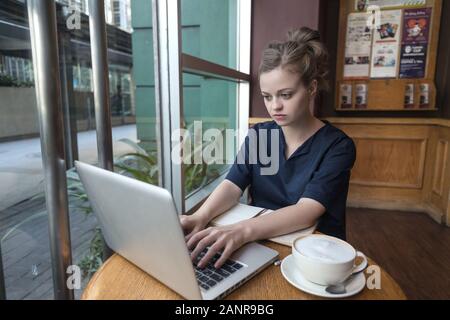 Sorridente giovane donna caucasica in un bar con computer portatile e caffè sul tavolo Foto Stock