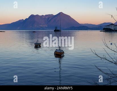 Paesaggio invernale sul Lago Maggiore con la luce soffusa del tramonto, barche a vela ormeggiata, ultimi raggi di sole sulle montagne riflessi nell'acqua Foto Stock