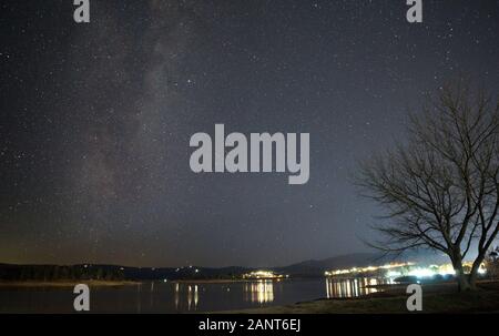 Via lattea la formazione di archi elettrici su un albero sfrondato sulla riva del lago Jindabyne con la città di Jindabyne in background Foto Stock