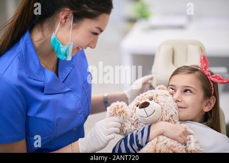 Bambina in un riunito dentale di nascondersi dietro il suo orsacchiotto Foto Stock