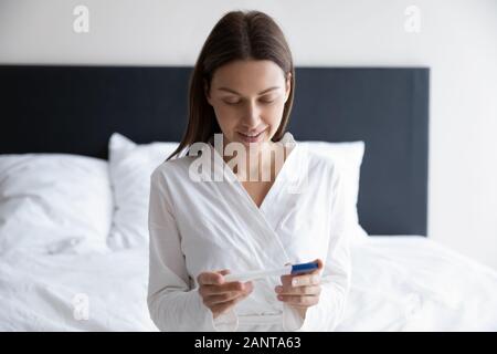 Donna sorridente seduta sul letto, guardando al test di gravidanza Foto Stock