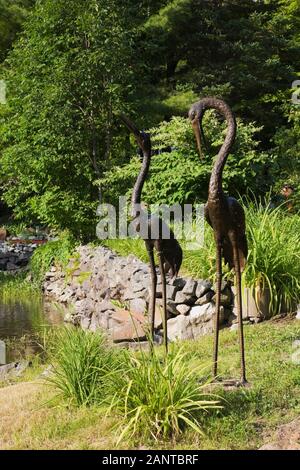 Due alte sculture di uccelli in metallo arone di bronzo sulla riva del fiume nel giardino di campagna di fronte cortile in estate Foto Stock
