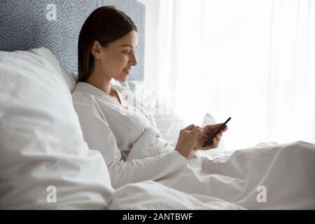 Sorridente giovane donna utilizzando phone, seduto sotto una coperta a letto Foto Stock